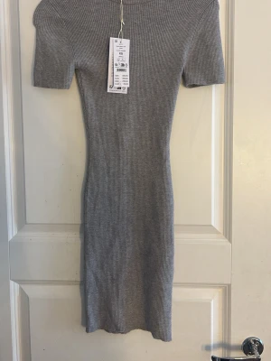 Grå ribbad kortklänning XS - Säljer en ljusgrå ribbad stickad klänning med kort ärm och rund hals. Modellen är figurnära och slutar ovanför knäna. Perfekt för dig som gillar en enkel men stilren look. Storlek XS.