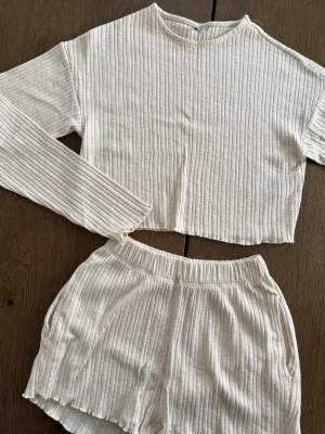Ribbat beige set från Nelly - Ribbat set från Nelly i beige. Setet består av en croppad långärmad topp och matchande shorts med resår i midjan. Jag har använt det som lounge/sov kläder. Storlek XS.