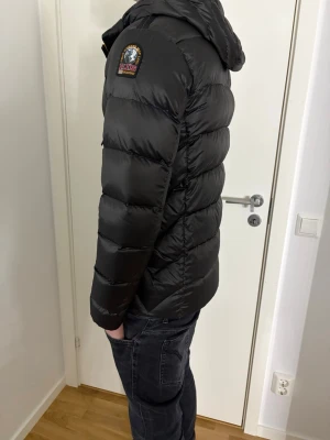 Svart pufferjacka från Parajumpers - Säljer en svart pufferjacka från Parajumpers, modell Sheen. Jackan har huva, flera fickor med dragkedja och tryckknappar samt en grön insida. Klassisk patch på ärmen och coola detaljer som orange flärp vid halsen.