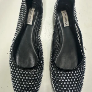 Steve Madden ballerina med strass stenar  - Svarta ballerinaskor från Steve Madden täckta med glittriga strassdetaljer. Skorna har rund tå, platt sula och meshpartier på sidorna för extra stil. Perfekta för dig som vill sticka ut med lite extra bling i vardagen.