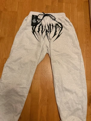 Grå mjukisbyxor BreatheDivinity  - Säljer ett par grå mjukisbyxor från Breathe Divinity med svart snörning i midjan och ett stort svart tryck framtill. Byxorna har avslappnad passform och resår i midjan för extra komfort. Perfekta för chill dagar eller streetwear-stil.