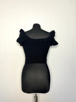 Svart stickad croppad topp H&M - Supersöt svart stickad topp från H&M med volangdetaljer längs den vida ringningen. Modellen är croppad och har korta ärmar som sitter offshoulder. Perfekt till höga jeans eller kjol för en trendig look.