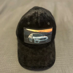 Custom keps med BMW motiv - Svart keps med mesh på baksidan och mjuk sammet framtill. Framsidan har en broderad patch med en vit BMW framför en solnedgång. Justerbar passform och coolt street-utseende, perfekt för bil intresserade .