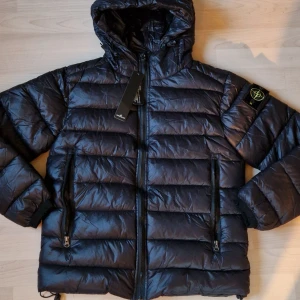 Svart pufferjacka från Stone Island - Snygg svart pufferjacka från Stone Island med huva och ikonisk patch på ärmen. Jackan har två stora dragkedjefickor fram, quiltad design och justerbar nederkant och luva. Perfekt för kalla dagar och har en cool streetwear-vibe. Storlek S! 🙌 Hör av er vid minsta fråga 🤍
