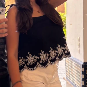 Svart linnetopp med vit broderi - Säljer en svart linnetopp med vit broderad blomdetalj längs den vågiga nederkanten. Toppen är ärmlös och har en rak halsringning med tunna axelband. Perfekt för varma dagar och ger en söt och fräsch vibe till din outfit.