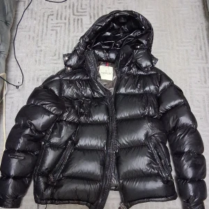 Svart Moncler pufferjacka med huva - Säljer en svart pufferjacka från Moncler med glansig finish och stor huva. Jackan har dragkedja framtill, två snedställda fickor med dragkedja och klassisk Moncler-logga på ärmen. Perfekt för kalla dagar när du vill hålla stilen.Passar i storlek S.