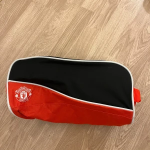 Manchester United röd och svart väska - Sportig väska i rött och svart med Manchester United-logga på framsidan. Tillverkad i 100% polyester och har dragkedja upptill. Perfekt för att bära träningskläder eller skor. Snygg kontrast med vit kant och praktiskt handtag på sidan.