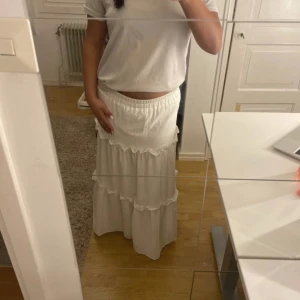 Vit volangkjol med resårmidja - Säljer en lång, vit kjol med flera volanglager och resår i midjan. Kjolen har en luftig och flowy känsla, perfekt för dig som gillar en romantisk och avslappnad stil. Passar snyggt med en enkel topp eller stickad tröja. Jag är ca 162 cm lång