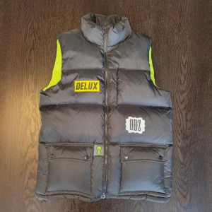 Svart dunväst DELUX ODZ, Signerad - Svart dunväst i storlek L från DELUX ODZ. Västen är inköpt på ODZs pop up store 2021 och är signerad på ryggen. Endast använd ett fåtal gånger. 