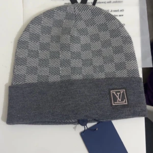 Grå L och V mössa  - Snygg grå mössa från L och V med klassiskt rutigt mönster och uppvikt kant. L och V-logga i grått framtill ger en lyxig touch. Tillverkad i mjuk ull som håller dig varm och stilren under kalla dagar.