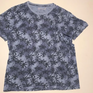 Blå t-shirt med palmmönster Polo Club - Snygg blå t-shirt från Polo Club med all-over palmmönster i mörkare nyanser. Klassisk rund halsringning och korta ärmar. Perfekt för en chill sommarlook.