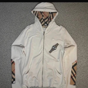 Vit hoodie från Burberry Brit - Snygg vit hoodie från Burberry Brit med dragkedja och klassiskt rutigt mönster i huvan och på ärmarna. Tröjan har huva, två fickor fram och subtilt broderad logga på bröstet. Perfekt för dig som gillar stilrena och exklusiva plagg.