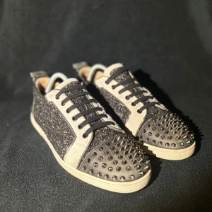Christian Louboutin skor  - Unika svarta sneakers från Christian Louboutin med glittrig textil, skinnpaneler och coola silverfärgade nitar på tån. Skorna har klassisk röd sula, snörning och en mix av material som ger en edgy vibe. Perfekta för dig som vill sticka ut med lyxig streetstyle.