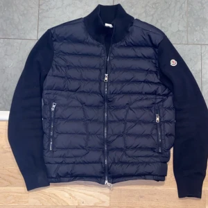 Moncler Cardigan - Moncler cardigan med quiltad front och stickade ärmar. Jackan har två dragkedjeförsedda fickor och en hög krage. Moncler-logga på vänster ärm. Perfekt för dig som vill ha en stilren och exklusiv look.