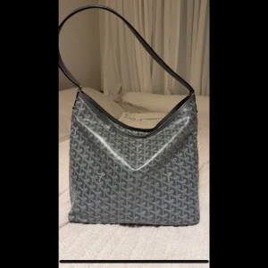 Grå axelväska  - Snygg grå axelväska med klassiskt mönster och lång axelrem. Väskan har en mjuk, fyrkantig form och är tillverkad i ett tåligt material med tryckt logotypmönster över hela ytan. Perfekt för dig som vill ha en stilren och ikonisk väska.