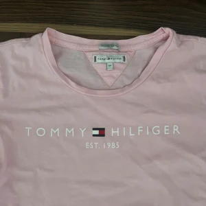 Rosa t-shirt från Tommy Hilfiger - Snygg ljusrosa t-shirt från Tommy Hilfiger med tryck framtill och klassisk logga. T-shirten är i mjuk bomull och har rund halsringning samt korta ärmar. Perfekt för en chill och stilren look.