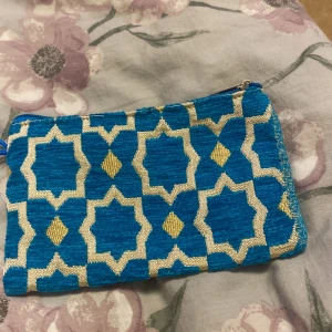 Blå och gul mönstrad plånbok - Snygg liten plånbok med orientaliskt inspirerat mönster i blått och gult. Väskan har en praktisk dragkedja upptill och är gjord i ett tygmaterial med glansiga detaljer. Perfekt för kort, mynt eller småsaker du vill ha med dig.