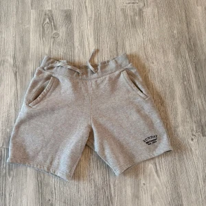 Grå sweatshorts från Sunday Sportswear - Grå mjuka sweatshorts från Sunday Sportswear med broderad logga på benet. Modellen har elastisk midja med snörning och två sidofickor. Perfekta för chill dagar eller träning. Tillverkade i bomullsmix för extra komfort.