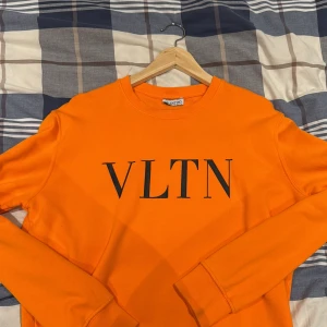 Orange sweatshirt från Valentino - Säljer en orange sweatshirt från Valentino med svart VLTN-tryck på bröstet. Tröjan har rund halsringning och långa ärmar. Perfekt för dig som vill sticka ut med en färgstark och trendig look.