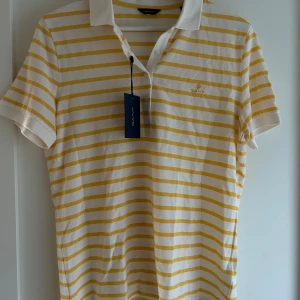 Vit och gul randig pikétröja GANT - Fräsch pikétröja från GANT i storlek M med vita och gula horisontella ränder. Klassisk krage, korta ärmar och knappar framtill. GANT-logga broderad på bröstet. Perfekt för dig som gillar stilrena och sportiga vibbar.