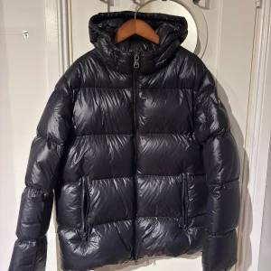 Svart Colmar pufferjacka med huva - Säljer en svart pufferjacka från Colmar Originals med glansig finish och huva. Jackan har två stora fickor med dragkedja, markerad logga på ärmen och hel dragkedja framtill. Perfekt för kalla dagar.