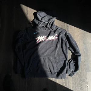 Svart Nike Miami hoodie - Svart hoodie från Nike med Miami-tryck i vitt, rosa och turkos framtill. Klassisk känguruficka och huva med snörning. Perfekt för dig som gillar streetwear och sportig stil.