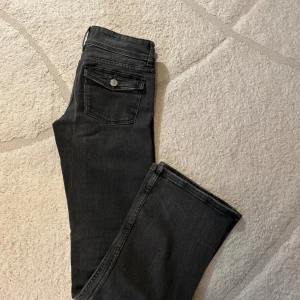 Svarta bootcut jeans med fickdetaljer - Snygga svarta jeans med bootcut passform och klassiska fickor med lock och knapp bak. Jeansen har låg midja och är tillverkade i mjukt denimtyg som sitter skönt. Perfekta för dig som gillar en tidlös look med lite extra edge. använda 3 ggr så som nya!!