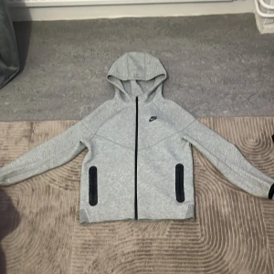 Grå NikeTech kofta - Snygg grå hoodie från Nike med svart dragkedja och svarta detaljer vid fickorna. Tröjan har huva, lång ärm och är tillverkad i mjuk bomullsmix. Tröjan har använts runt 5-10 gånger och har inga synliga fläckar eller något vidare! Storleken är XL i barnstorlek 158-170cm Pris kan diskuteras!
