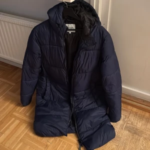 Mörkblå lång pufferjacka med huva - Säljer en mörkblå pufferjacka från Zara med huva och långa ärmar. Jackan är quiltad och har dragkedja framtill. Den är lång i modellen och har två sidofickor. Perfekt för kalla dagar när du vill hålla dig varm och ändå se chill ut.