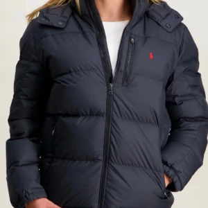 Svart pufferjacka från Polo Ralph Lauren - Svart pufferjacka från Polo Ralph Lauren med röd logga. Jackan har huva, flera dragkedjefickor och quiltad design. Materialet är syntet och polyester. Pris kan diskuteras vid snabbt köp.