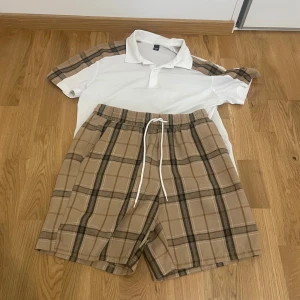 Beige rutiga shorts med snörning - Säljer ett par beige rutiga shorts från Manfinity i storlek M. Shortsen har elastisk midja med vit snörning och ett avslappnat fit. Perfekta för sommardagar och chill häng. Mjukt material och klassiskt rutmönster i beige och bruna toner.