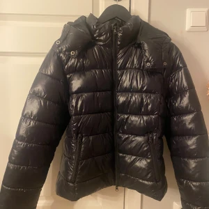 Svart pufferjacka från 157, M - Svart pufferjacka från 157 i storlek M. Jackan har en glansig finish, huva och två fickor med dragkedja. Den är quiltad med breda paneler och har hel dragkedja framtill. Perfekt för kalla dagar och har en modern, 