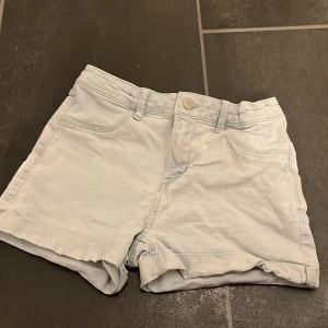 Ljusblå jeansshorts från H&M - Snygga ljusblå jeansshorts från H&M med klassisk femficksdesign och uppvikta benslut. Dessa shorts är också väldigt lätta att spänna in vid midjan med hjälp av knappar. De har en fin guldig dragkedja och en silvrig knapp. Har andvändts mycket men ända väldigt fina och kommer passa perfekt till sommarn 2026.😊