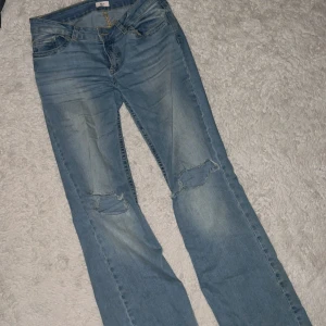 Ljusblå jeans med slitna knän från Nelly - Säljer ett par ljusblå jeans från Nelly med snygga slitningar på båda knäna och dekorativa nitar på bakfickan. Klassisk femficksmodell med gylf och knapp, samt guldfärgade detaljer. Perfekt för dig som gillar en avslappnad och trendig look.