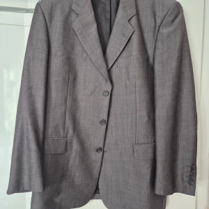 Kavaj - Snygg grå kavaj från Uptown Classic i storlek 52. Klassisk modell med tre knappar framtill, två fickor med lock och en bröstficka. Kavajen har en stilren, rak passform och är tillverkad i ett mjukt material som ger en elegant look.