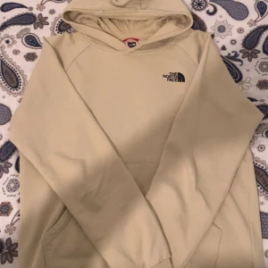 Beige hoodie från The North Face - Säljer en beige hoodie från The North Face med stor logga och texten 'Never Stop Exploring' på ryggen. Tröjan har huva, långärmad design och en klassisk känguruficka framtill. Perfekt för chill dagar och streetwear-stil.