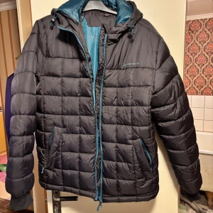 Svart pufferjacka med blå detaljer - Svart pufferjacka från 4F med blå dragkedjor och foder. Jackan har huva med snörning, två sidofickor och quiltad design. Perfekt för kalla dagar och har en modern, sportig look. Passar dig som gillar stil och funktion.