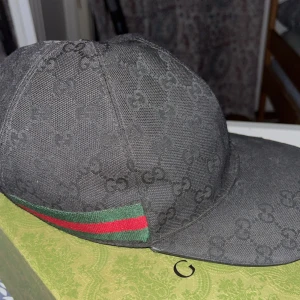 Svart Gucci keps XL - Snygg svart keps från Gucci i storlek XL med klassiskt GG-monogram över hela kepsen. På sidan finns den ikoniska gröna och röda randen. Materialet är slitstark bomull och kepsen har en böjd skärm. Perfekt för dig som vill ha en lyxig streetstyle-look.