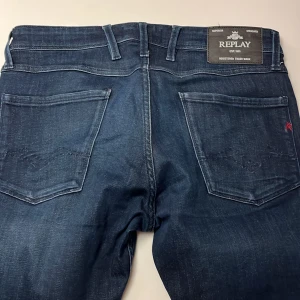 Replay Anbass jeans mörkblå herr - Snygga mörkblå Replay Anbass jeans med klassisk femficksdesign och dragkedjegylf. Jeansen har diskreta slitningar och en stilren look, perfekta för dig som gillar en modern och enkel stil. Märkespatch bak i midjan och detaljerad logga på fickan.