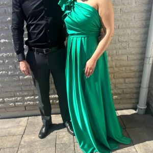 Grön långklänning med oneshoulder - Säljer en elegant grön långklänning med oneshoulder och volangdetalj vid axeln. Klänningen har ett svepande fall och markerad midja, perfekt för dig som vill sticka ut med en färgstark look. Materialet är i satin  och känns lyxigt mot huden. Går att sy in eller sy ut samt korta ner.  Inköps pris 5000 säljer 3000 finns i Göteborg och Jönköping för upphämtning 