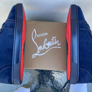 Mörkblå sneakers från Christian Louboutin - Snygga mörkblå sneakers från Christian Louboutin i mocka med ikonisk röd sula och broderad logga på hälen. Skorna har rund tå, snörning och exklusiv känsla med beige innersula. Perfekta för dig som vill sticka ut med lyxiga detaljer.