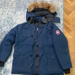 Canada Goose Jacka - Säljer en blå Canada Goose jacka med äkta päls på huvan och klassisk logga på ärmen. Jackan har flera stora fickor, dragkedja och knappar framtill samt ribbade muddar. Perfekt för kalla dagar och har en snygg, tidlös look. Priset är förhandlingsbart vid snabb affär