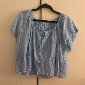 Ljusblå blus från Scotch & Soda - Supersöt ljusblå blus från Scotch & Soda med broderade cirklar, kort ärm och knytning vid halsen. Blusen har en luftig och flowy passform, perfekt för varma dagar. Snygg volangdetalj nedtill och lätt strukturerat tyg. Ny utan prislapp.