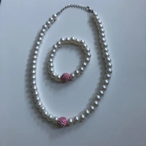 Pärlset halsband & armband med rosa detalj - Säljer ett matchande set med halsband och armband av vita pärlor. Båda har en rund rosa pärla med glittrande stenar som snygg detalj. Halsbandet har justerbart spänne och armbandet är elastiskt. Perfekt för dig som gillar klassisk stil med en twist.