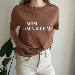 Brun t-shirt „I Am A Rich Man“ Small Tits Club - Brun t-shirt i bomull från Small Tits Club med vit text framtill: 'MOM, I AM A RICH MAN'. Klassisk rund halsringning och normal/oversized passform. 
