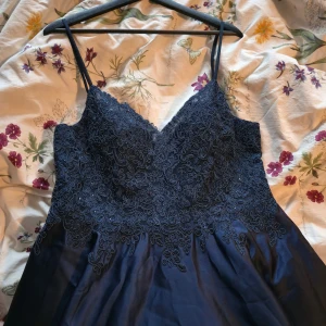 Mörkblå långklänning från JJ's House - Oanvänd elegant mörkblå långklänning från JJ's House med smala axelband, v-ringad spetsliv med broderade blommor och glittriga detaljer. Kjolen är i glansigt tyg som faller snyggt och har två fickor. Perfekt för bal eller festliga tillfällen. Den är uppsydd men borde vara lätt att sprätta upp. Finns i Östersund, kan tas till Ragunda kommun också.