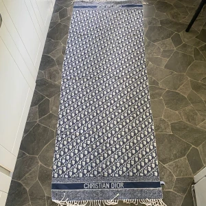 Blåvit halsduk från Christian Dior - Snygg blåvit halsduk från Christian Dior med klassiskt logomönster över hela ytan och fransar i ändarna. Halsduken har en rektangulär form och tydlig logga längs ena kanten. Perfekt accessoar för att lyfta din outfit.