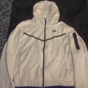 Vit Nike teech med huva - Snygg vit vindjacka från Nike med svart logga på bröstet och svarta detaljer längs dragkedjan och ärmen. Jackan har huva, hel dragkedja och en extra ficka med dragkedja på ena ärmen. Perfekt för dig som gillar sportig stil. Pris går att diskuteras 