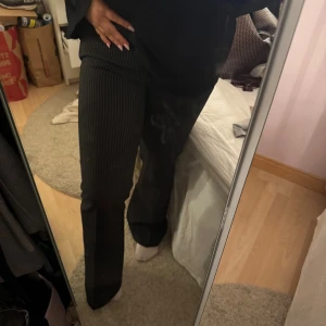 Svarta bootcut kostymbyxor med ränder - Säljer ett par svarta kostymbyxor med tunna vita ränder. Byxorna har bootcut-modell och sitter snyggt i midjan. Perfekta för dig som gillar klassisk stil med en twist. Materialet känns mjukt och följsamt.
