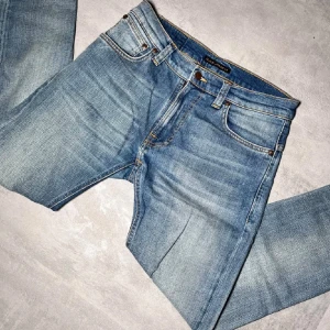Nudie Jeans  - Säljer ett par klassiska blå jeans från Nudie Jeans i storlek 30/30. Jeansen har fem fickor, orangea sömmar och Nudie-loggan på bakfickorna. Snygg tvättad look och raka ben. Perfekta för dig som gillar stilrena och tidlösa jeans.